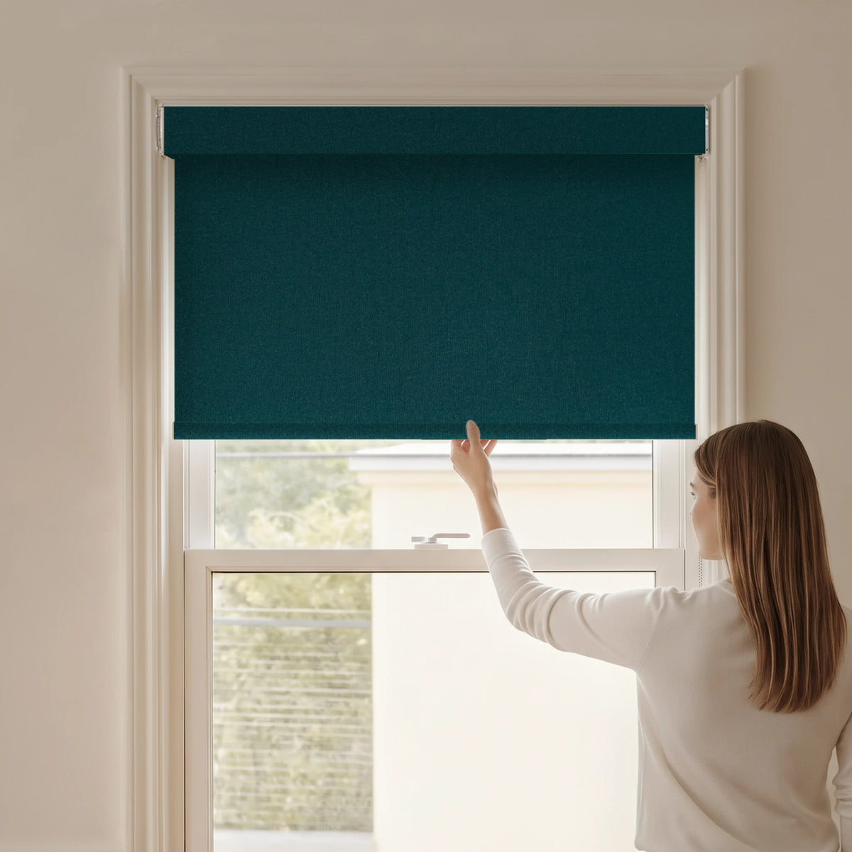 Woman adjusting a Graywind blackout manual-motorized roller shade in Plush Matte (Blue) fabric.