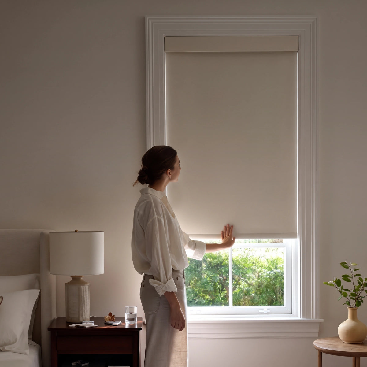 Woman adjusting a Graywind manual-motorized roller shade by hand in a bedroom.
