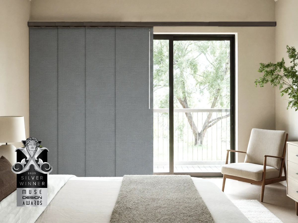 A modern bedroom interior showing a large gray sliding wardrobe door next to a glass sliding door with a view of trees outside. A beige armchair, white bed, and a silver award badge are also visible.