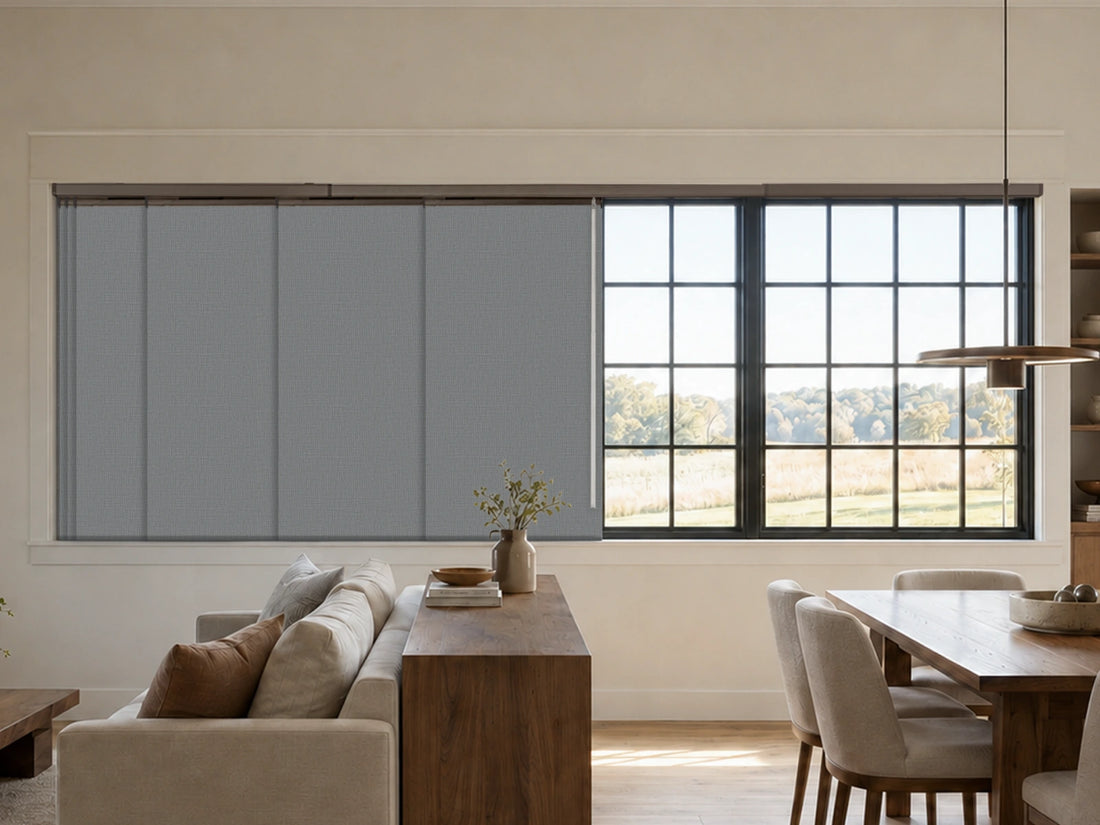 A modern interior room showing a large window with gray roller blinds partially drawn, alongside a dining table, chairs, sofa, and sideboard in a bright, contemporary space.