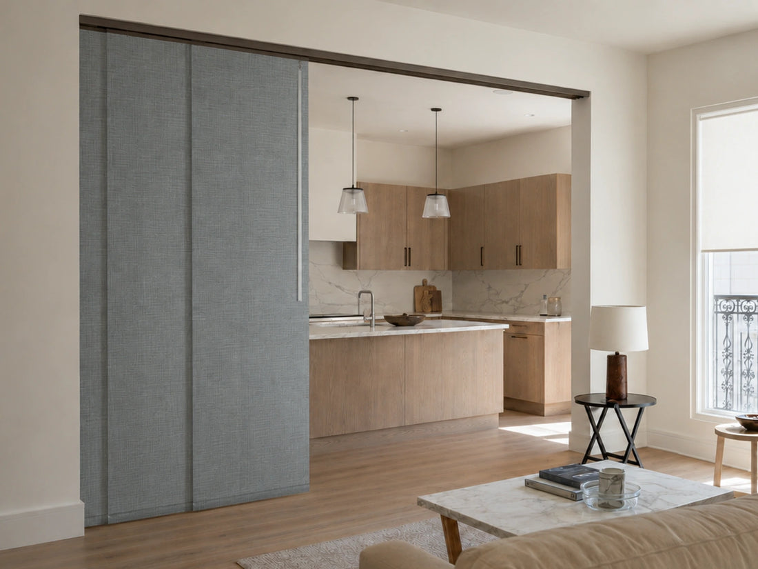 A modern interior room showing a large gray fabric-covered sliding door or cabinet panel on the left, with a visible kitchen in the background and a living area with a coffee table and lamp on the right.