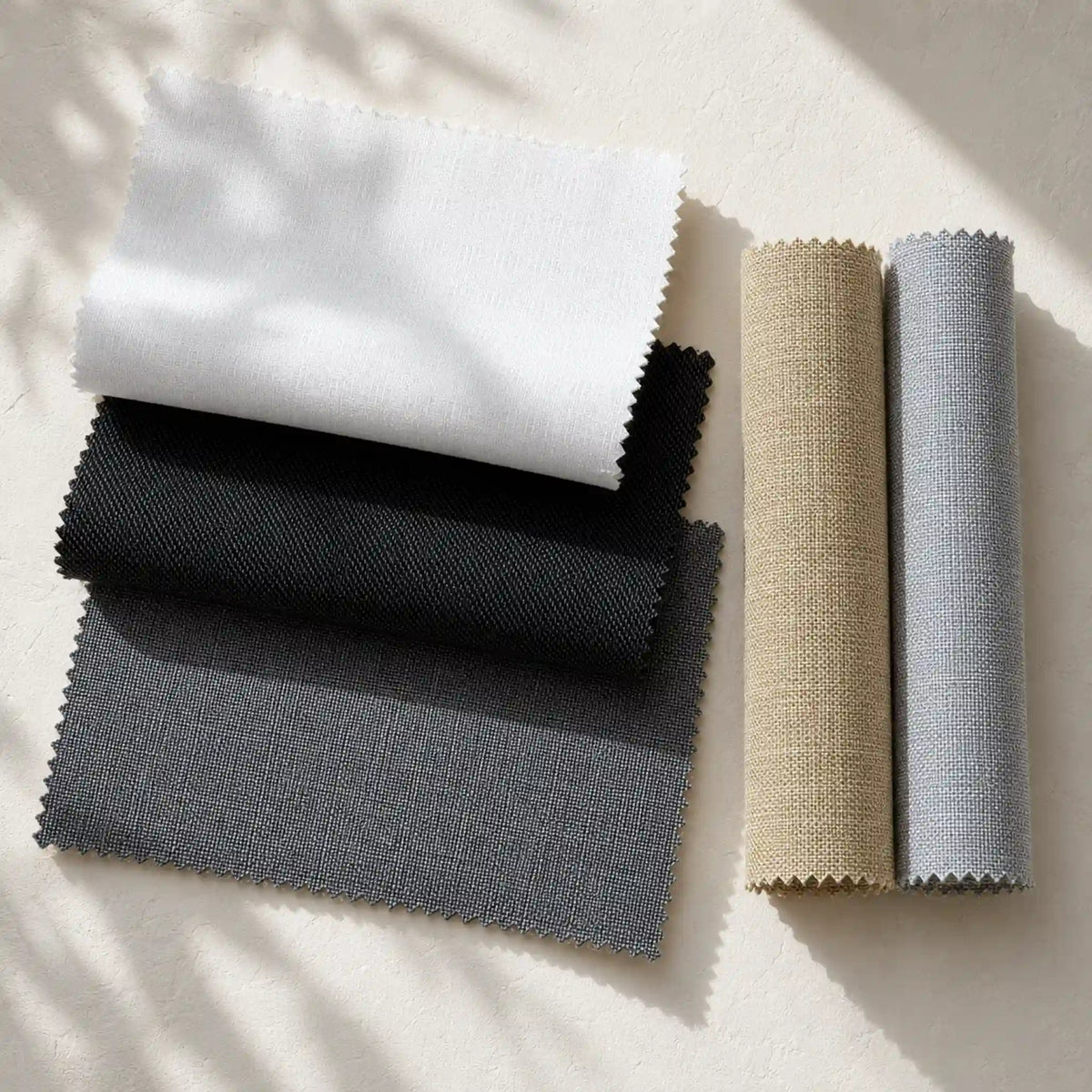 Four fabric color swatches with scalloped edges are arranged on a light beige surface with soft leaf shadows. Two swatches are folded fabric pieces in white, black, and dark gray, and two are rolled fabric pieces in beige and light gray.