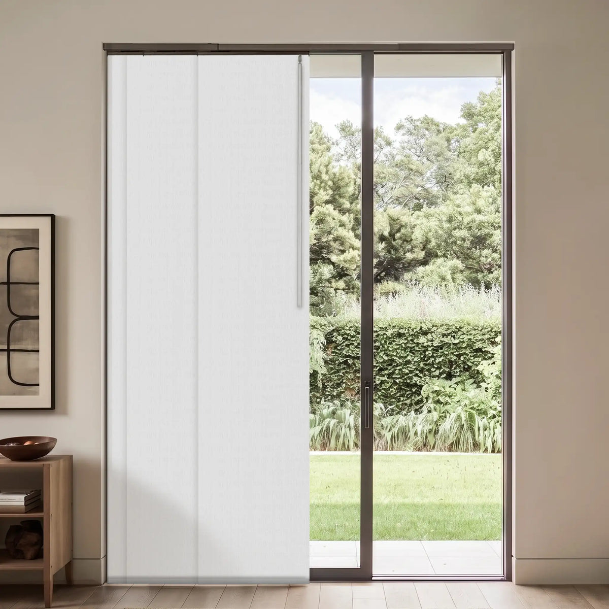 A modern sliding door system with two white vertical panels and a dark metal frame, set in a minimalist beige room with a view of a green garden outside.