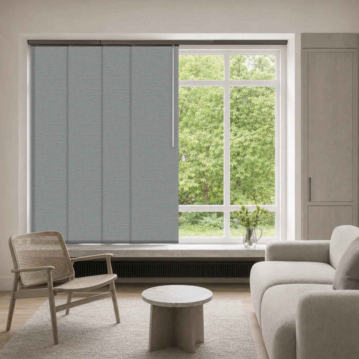 A modern living room interior with a light gray textured roller blind partially drawn over a large window, with a white sofa, woven armchair, and small round table in front of the window.