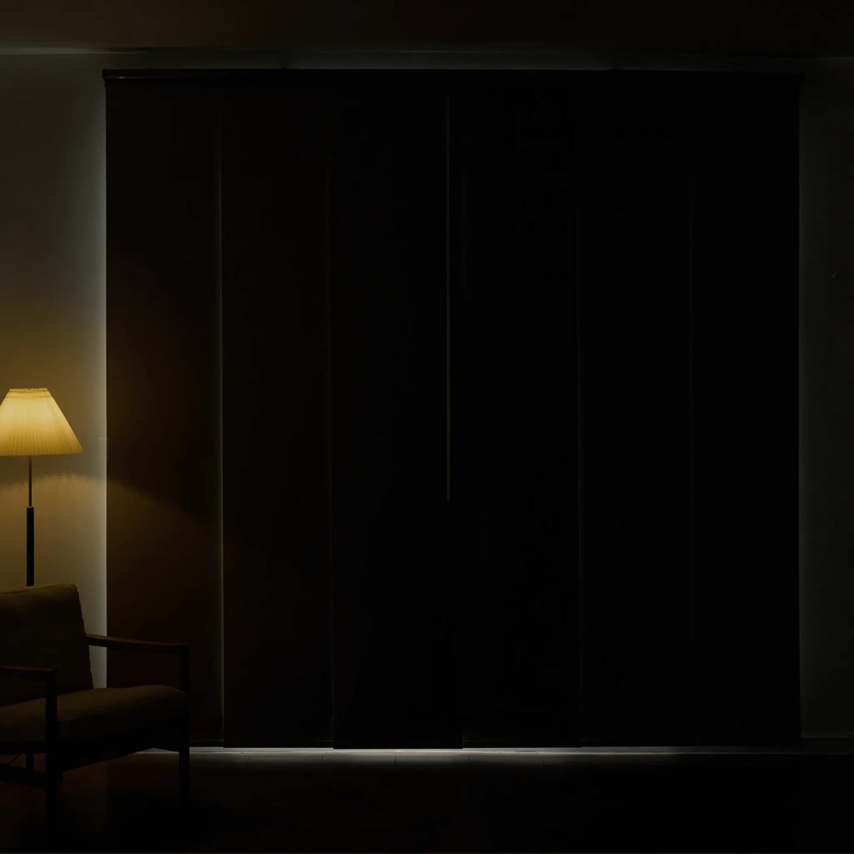 A set of dark, nearly black sliding or folding doors in a dimly lit room, with a warm floor lamp glowing on the left and a faint light strip along the bottom.