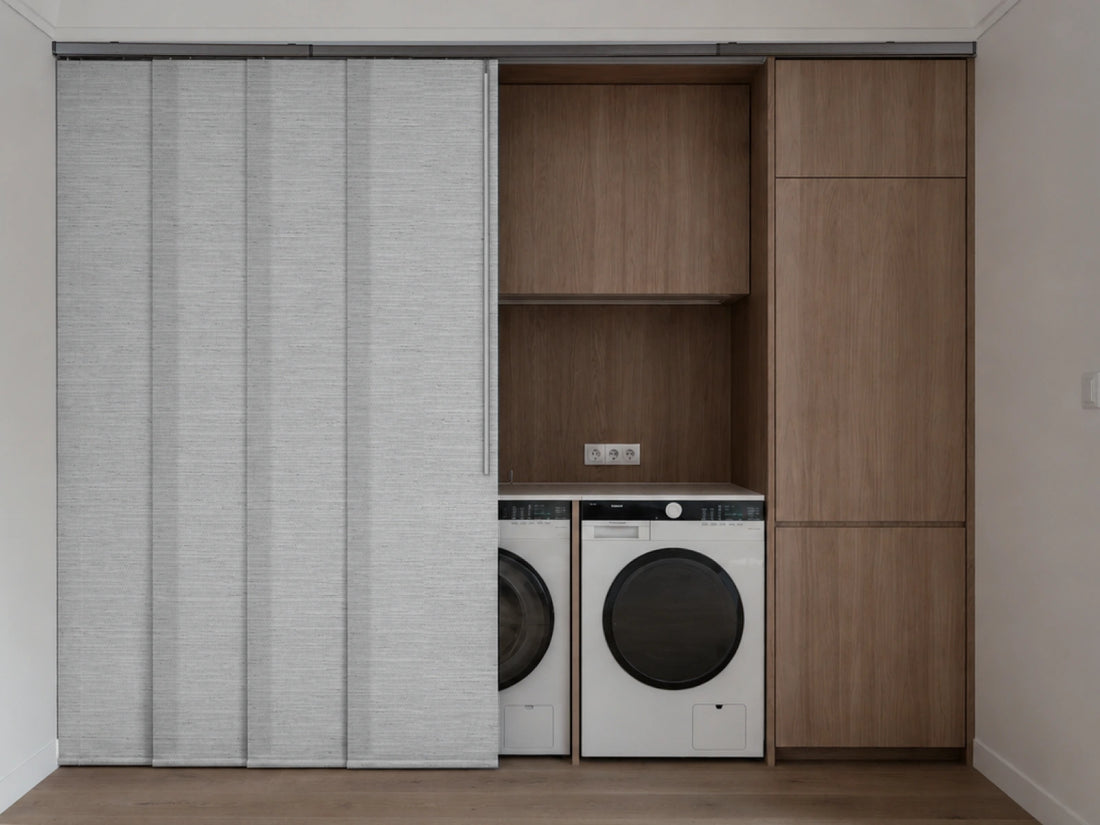 A modern built-in laundry nook with a sliding light gray textured door on the left, revealing a white and black front-loading washing machine and a white dryer beneath open wood shelving, next to a tall wood cabinet.