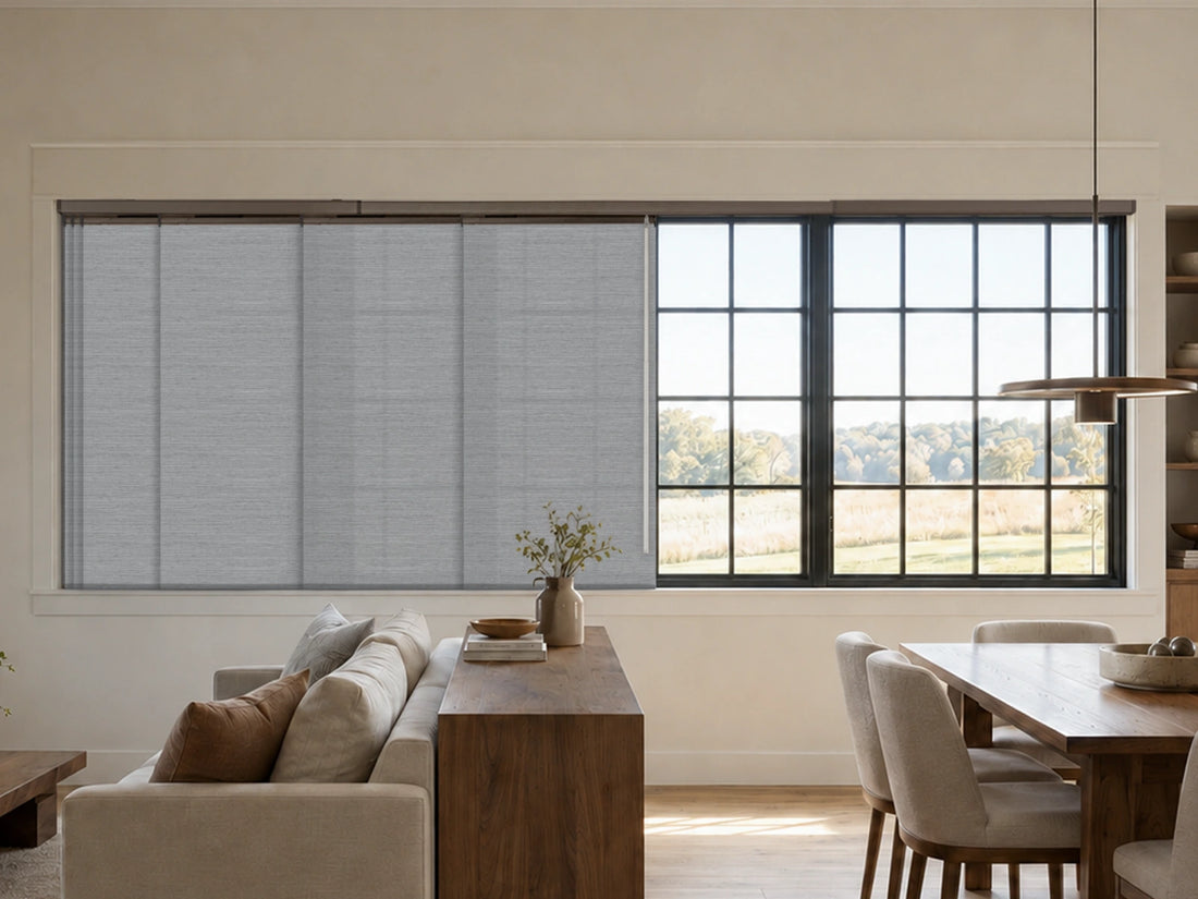A modern interior room showing a large window treatment setup with light gray roller shades on the left and black-framed multi-pane windows on the right, overlooking an outdoor landscape. A sofa, wooden console, dining table, and pendant light are visible in the room.