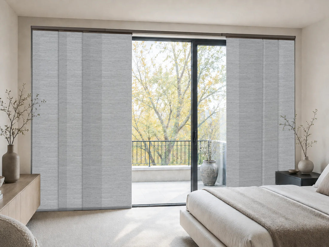 Light gray textured roller blind panels installed on a large sliding glass door in a modern, sunlit bedroom.