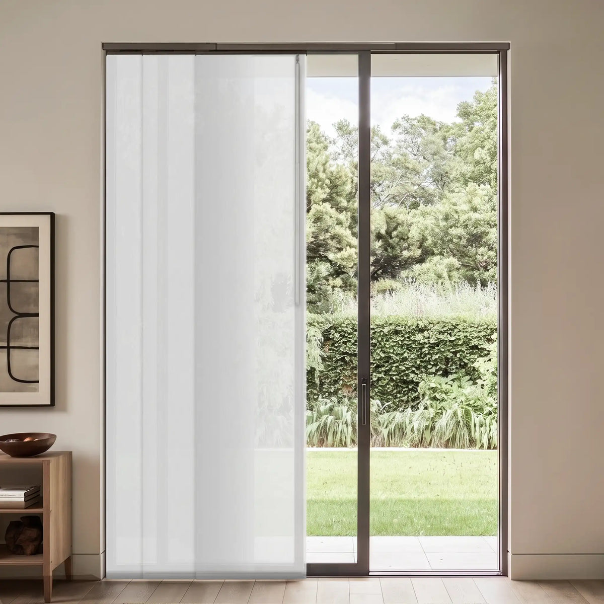 A white sheer vertical sliding door panel installed in a dark-framed sliding glass door, showing a green garden outside, set in a modern minimalist room.