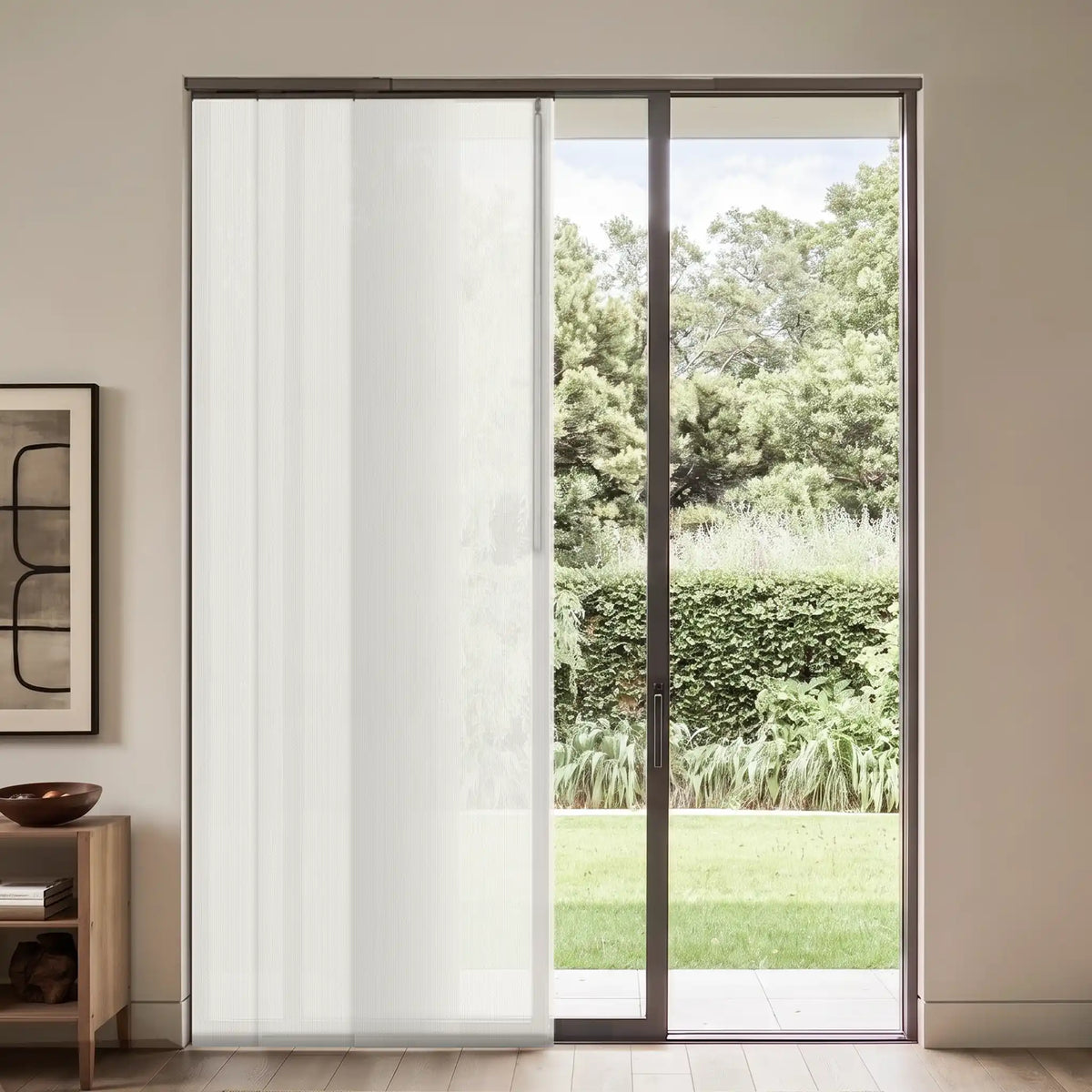 A modern sliding glass door with a semi-sheer white vertical blind panel on the left, set in a minimalist room with a wooden side table and framed wall art, looking out onto a green garden.
