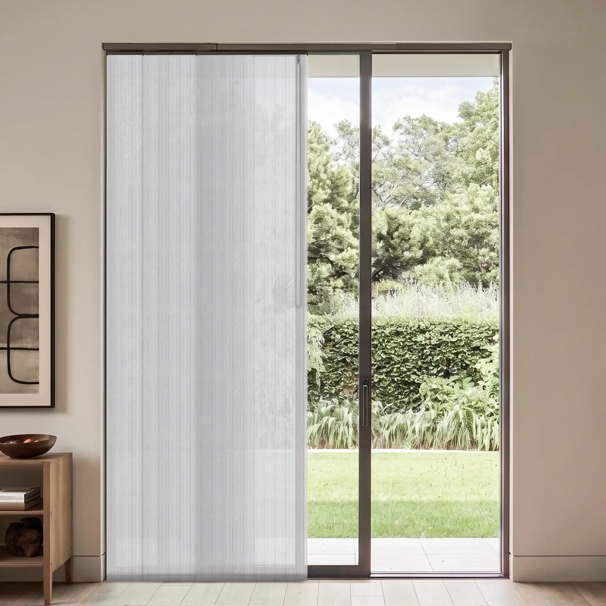 A white vertical blind roller shade partially drawn over a large sliding glass door, set in a modern room with a garden view.