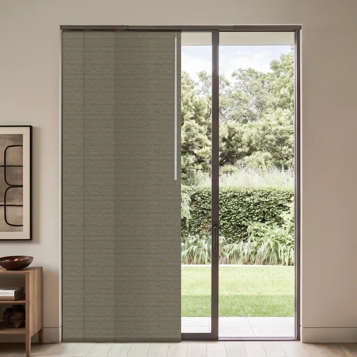 A modern sliding glass door with a textured gray roller blind partially drawn on the left, set in a minimalist room with a wooden side table and framed wall art.