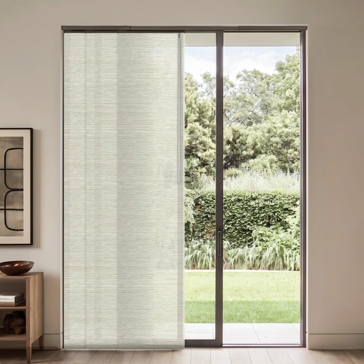 A light beige textured roller blind partially drawn over a sliding glass door, showing a green garden outside, in a modern room setting.