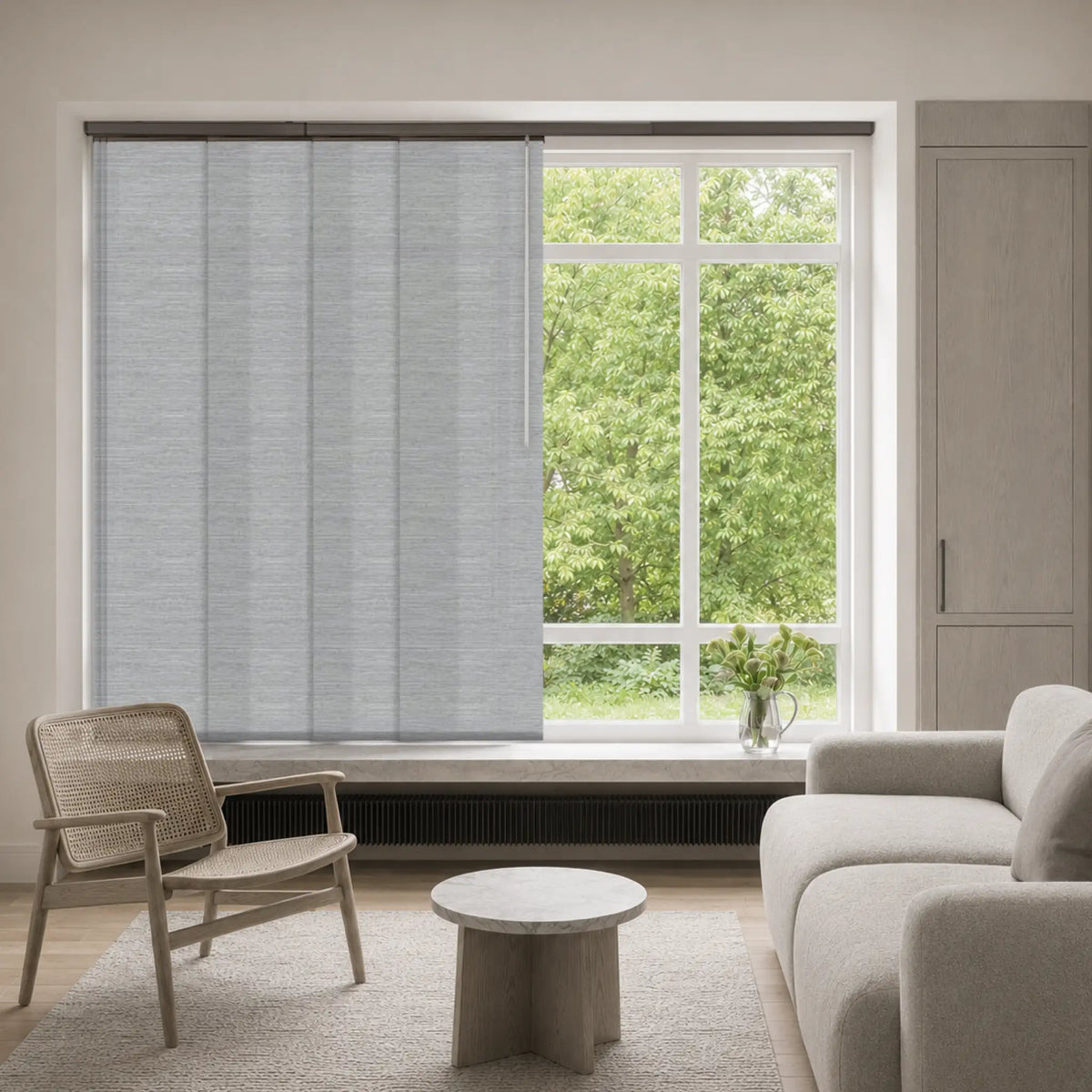 Light gray textured vertical roller blinds partially drawn over a large window in a modern, neutral-toned living room with a chair, sofa, and side table.