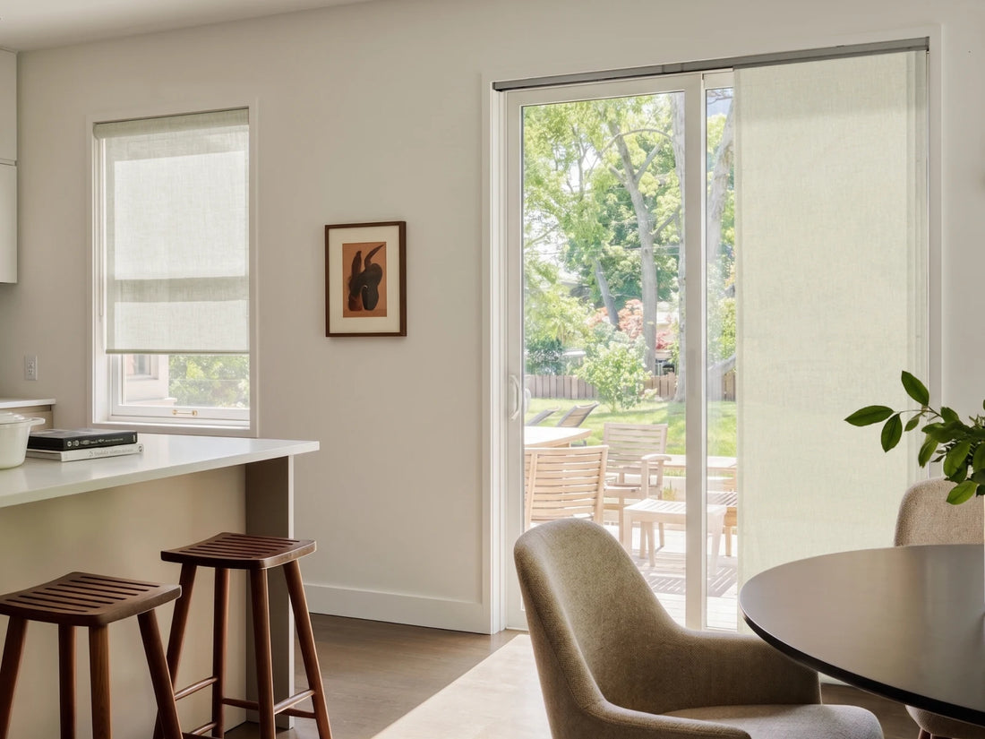 A set of light beige roller blind curtains shown installed on a sliding glass door and a smaller window in a modern, sunlit kitchen and dining room.