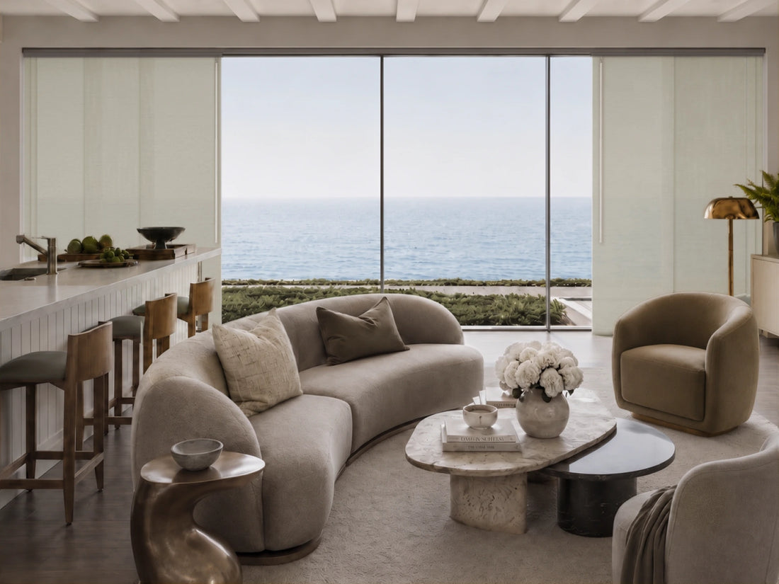 A modern living room with a curved beige sofa, a matching accent chair, and a stone coffee table, set against a large window overlooking the ocean.