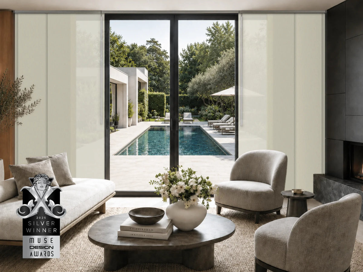 A modern living room with large floor-to-ceiling glass doors overlooking a swimming pool and garden. The room contains light beige upholstered armchairs, a sofa, a round coffee table with books and a vase of flowers, and a dark fireplace on the right.