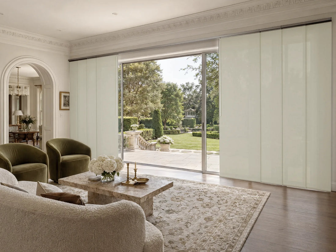 A set of modern, floor-to-ceiling sliding glass doors with light beige/cream panels, shown partially open in a luxurious living room with hardwood floors, a stone coffee table, and a garden view.