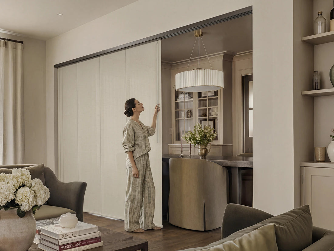 A woman in a matching beige lounge set stands in a modern living room, reaching toward a large sliding door that opens into a kitchen area. The room features neutral beige walls, a round pendant light, and tasteful home decor.