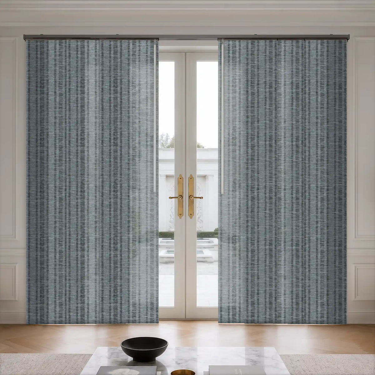 A pair of tall window curtains with a textured vertical stripe pattern in slate blue and gray, hanging on a rod over a set of white French doors in a bright room.