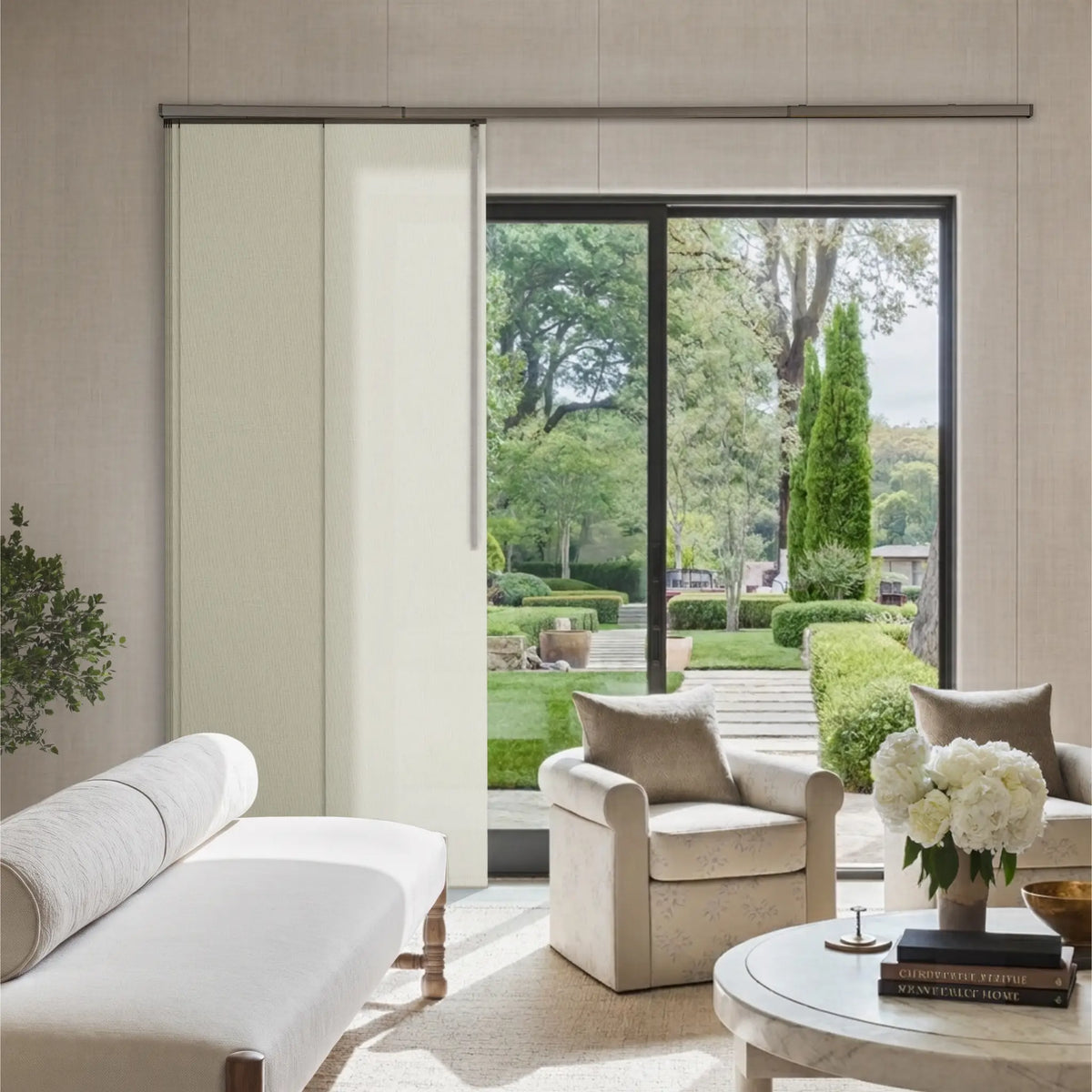 A modern living room interior with a large sliding glass door opening to a landscaped garden. The room features light beige upholstery, including a chaise lounge, armchair, and matching cushions, with a round white coffee table holding a vase of white flowers.