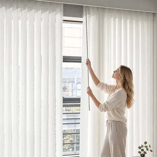 A woman in a cream-colored outfit adjusts a long metal rod inside a pair of white sheer  vertical blind hanging over a window.