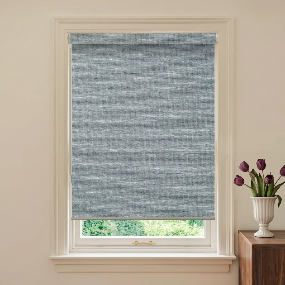 Window with a blue roller blind and a vase of purple tulips on a wooden side table.