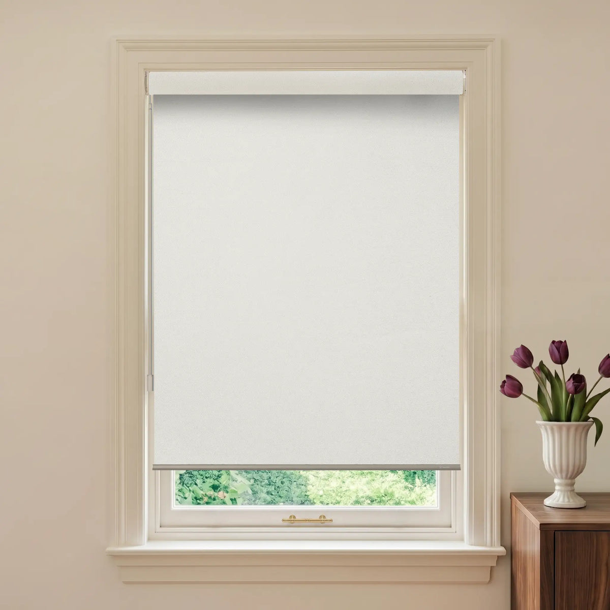 Window with white roller blind and a vase of purple tulips on a wooden cabinet.