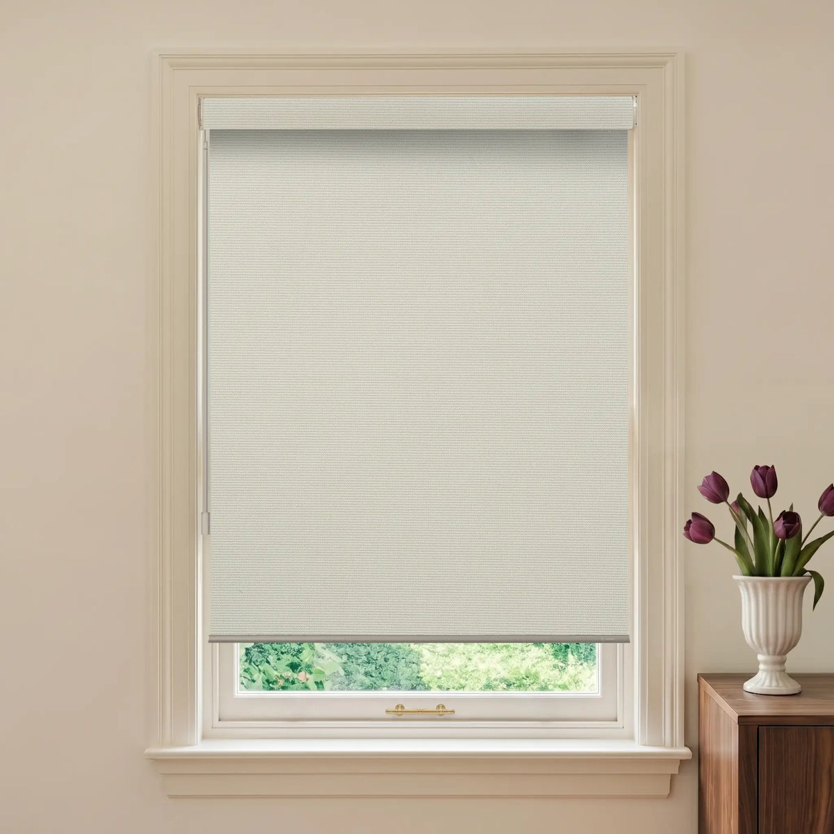 Window with beige roller blind and a vase of purple tulips on a wooden cabinet.