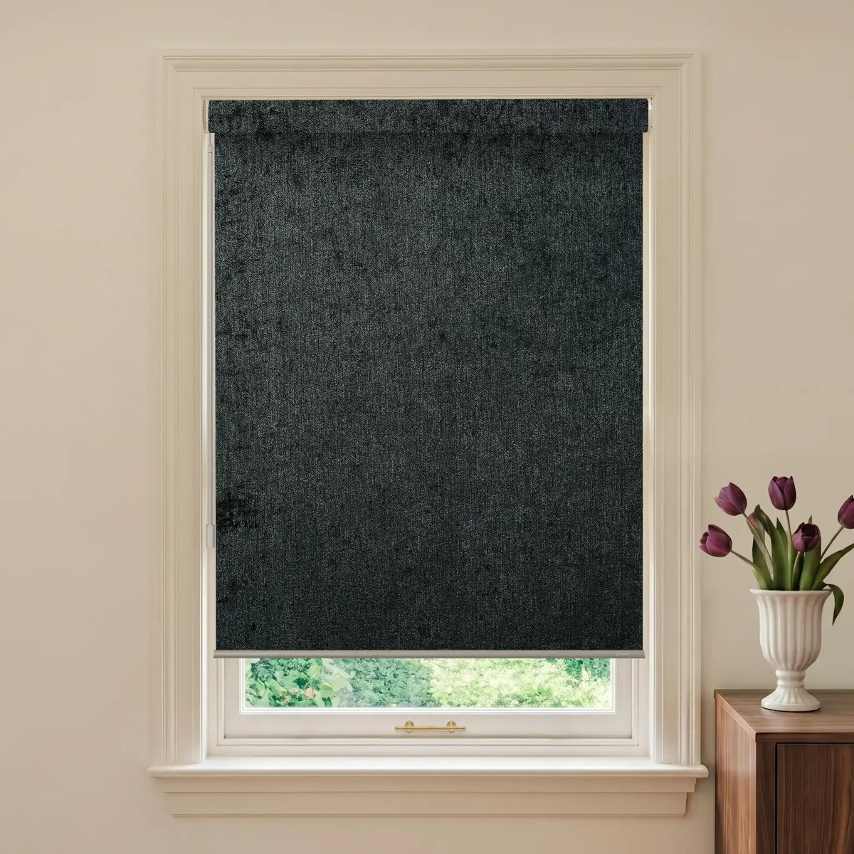 Black roller blind on a window with a vase of flowers on a table.
