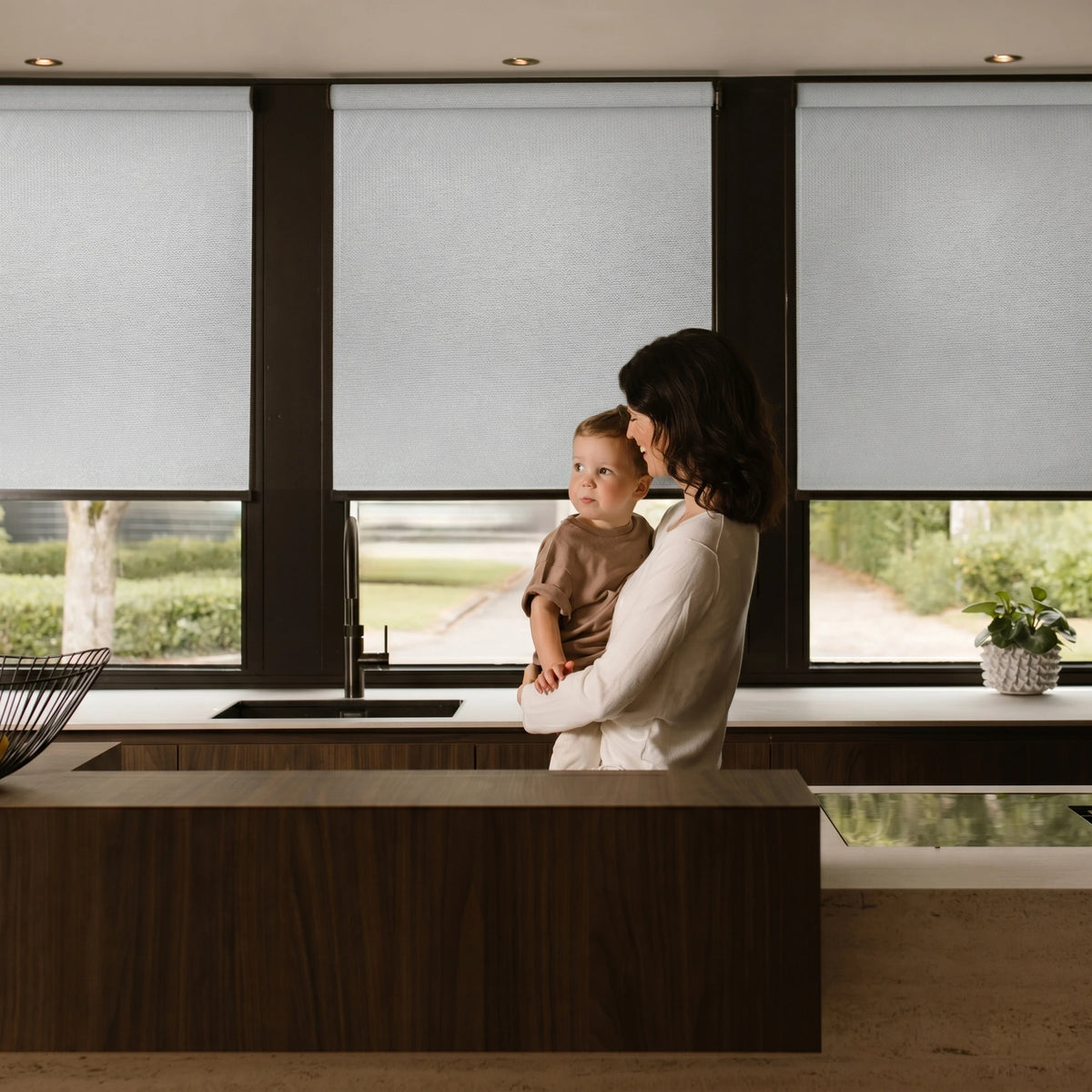Woman holding a child in a modern kitchen with large windows treatment.