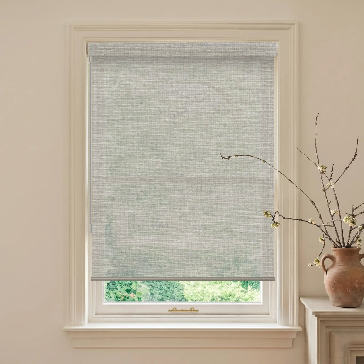 Window with a light gray roller blind, beige walls, and a vase with branches on a windowsill.