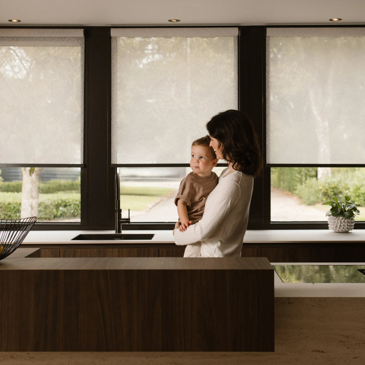 Woman holding a child in a modern kitchen with large graywind roller shades.