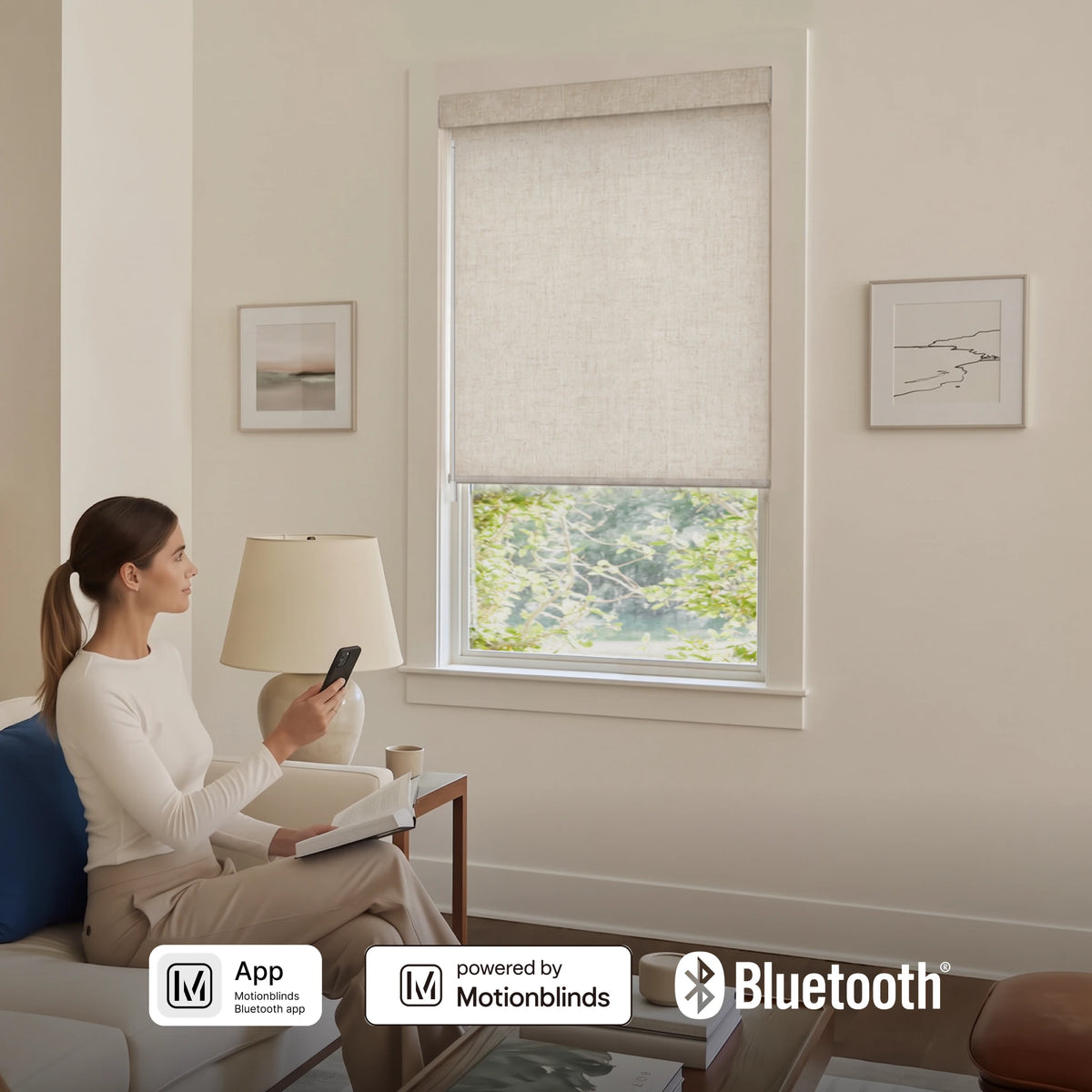 A woman sits on a sofa in a living room, using a phone to control a window blind. The blind is a light beige linen-style roller shade with a matching fabric panel, shown in a real interior setting with framed wall art, a lamp, and a coffee table.