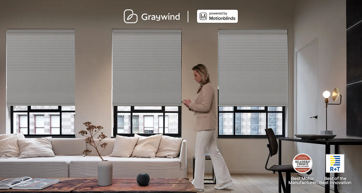 Woman standing in a modern living room with motorized cellular shades, featuring Graywind branding and Motionblinds branding.