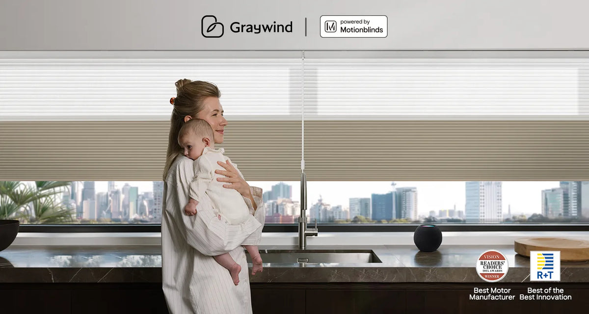 Woman holding a baby in a modern kitchen with day and night blinds and cityscape view, featuring Graywind branding and Motionblinds branding.