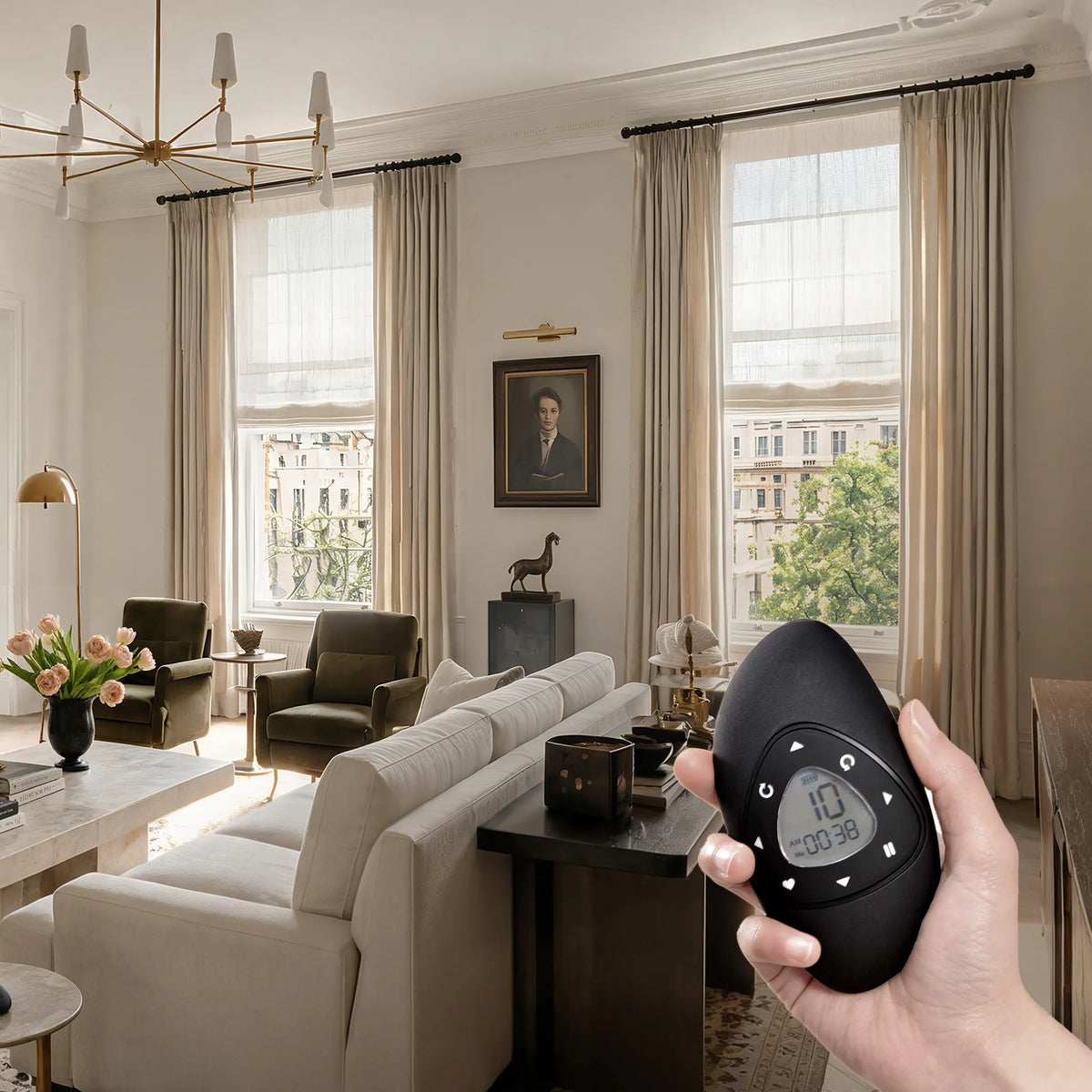 A hand holds a black remote control in a modern, elegantly furnished living room with beige curtains, cream sofas, and large windows.