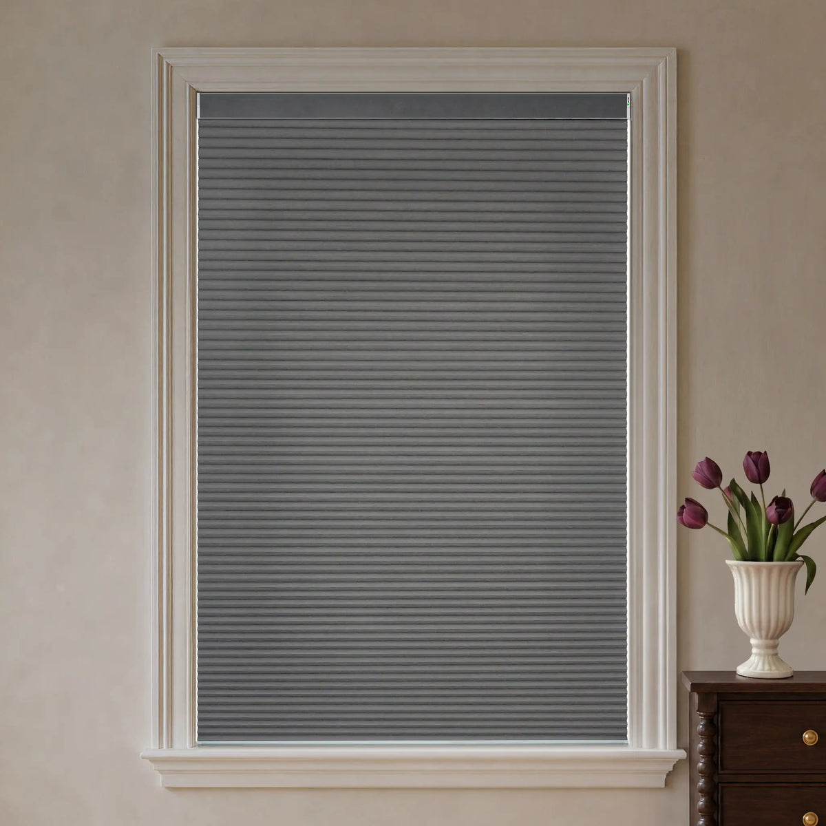 A gray cellular window shade installed in a white-framed window, shown in a styled interior setting with a vase of purple tulips on a dark wood side table.