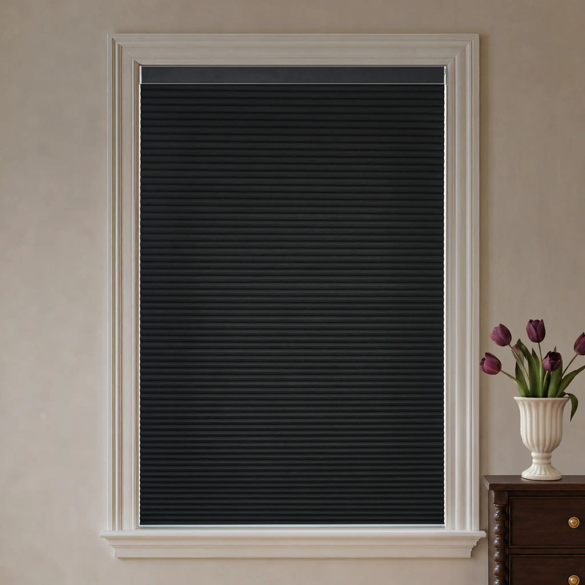 Black cellular window shade installed in a white-trimmed window, shown against a beige wall with a small vase of purple tulips on a wooden table at the right.