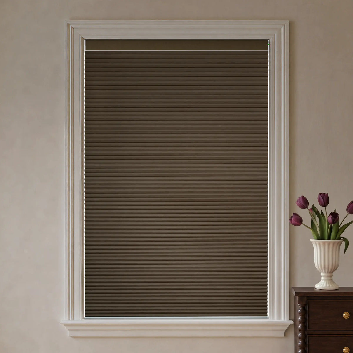 A dark brown cellular window shade installed in a white-trimmed window, shown in a styled room setting with a vase of purple tulips on a wooden cabinet.