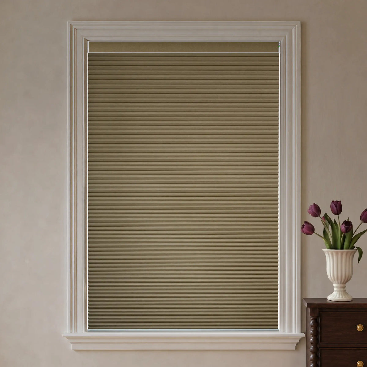 A beige cellular window shade installed in a white window frame, shown in a styled room setting with a vase of purple tulips on a wooden cabinet to the right.