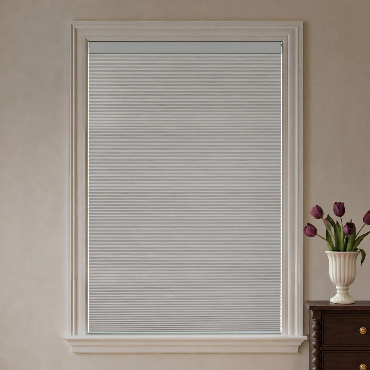 A Light Grey cellular window shade installed in a white window frame, shown against a beige wall with a small wooden cabinet and a vase of purple tulips visible on the right.