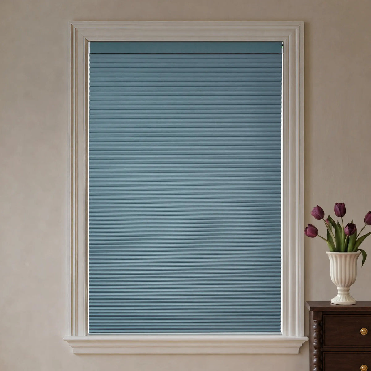 A blue-gray cellular window shade installed in a white-framed window, shown in a styled room setting with a vase of purple tulips on a dark wood table to the right.