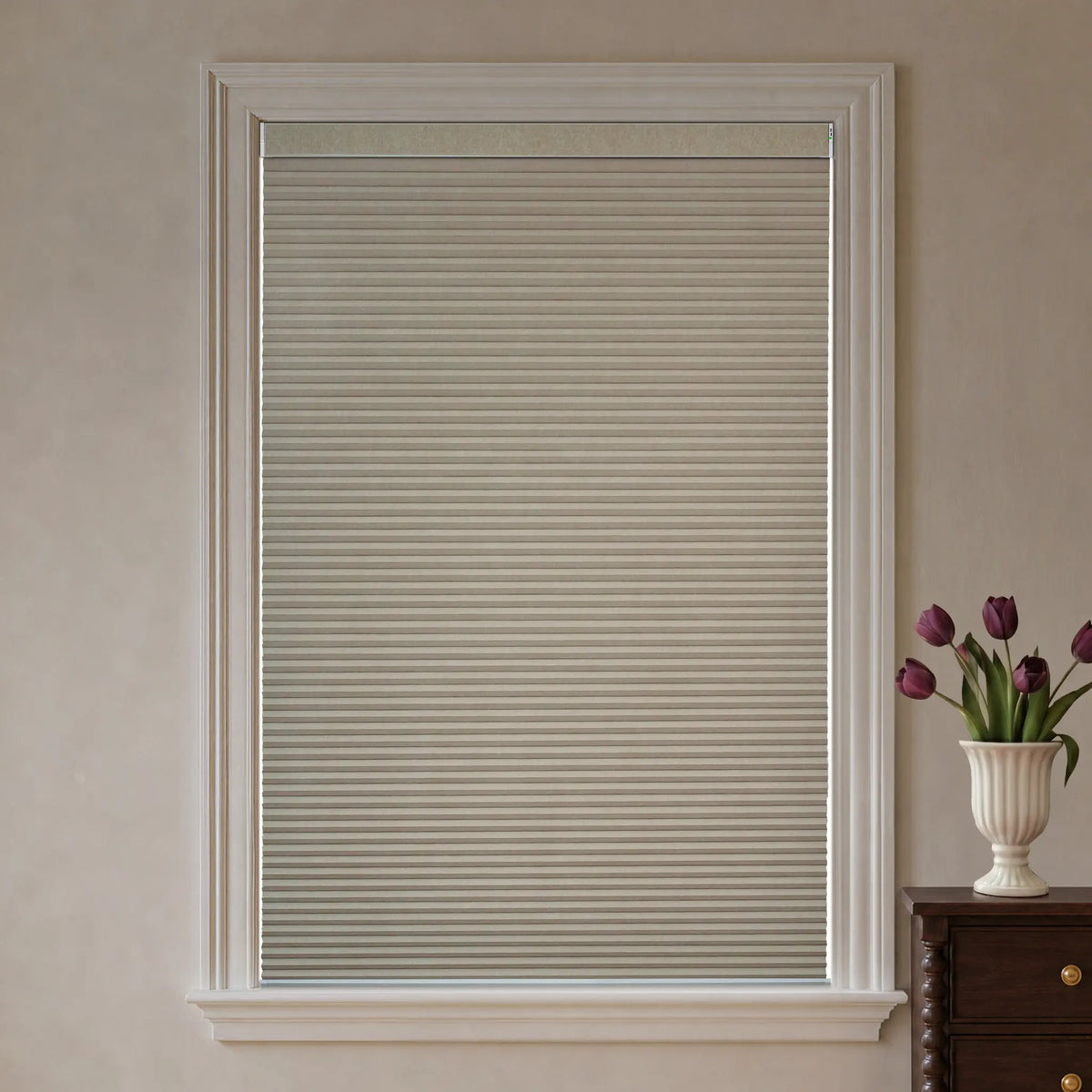 A coffee cellular window shade installed in a white-trimmed window, shown in a styled room setting with a vase of purple tulips on a dark wood chest.