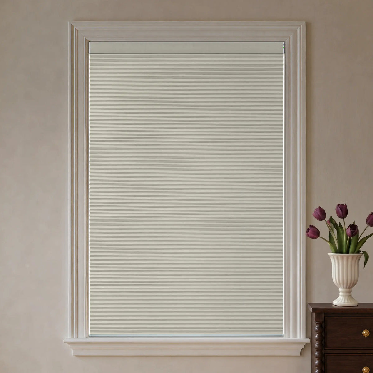 Beige motorized blackout cellular shades installed in a white window frame, shown on a beige wall with a dark wood cabinet and a vase of purple tulips visible on the right.