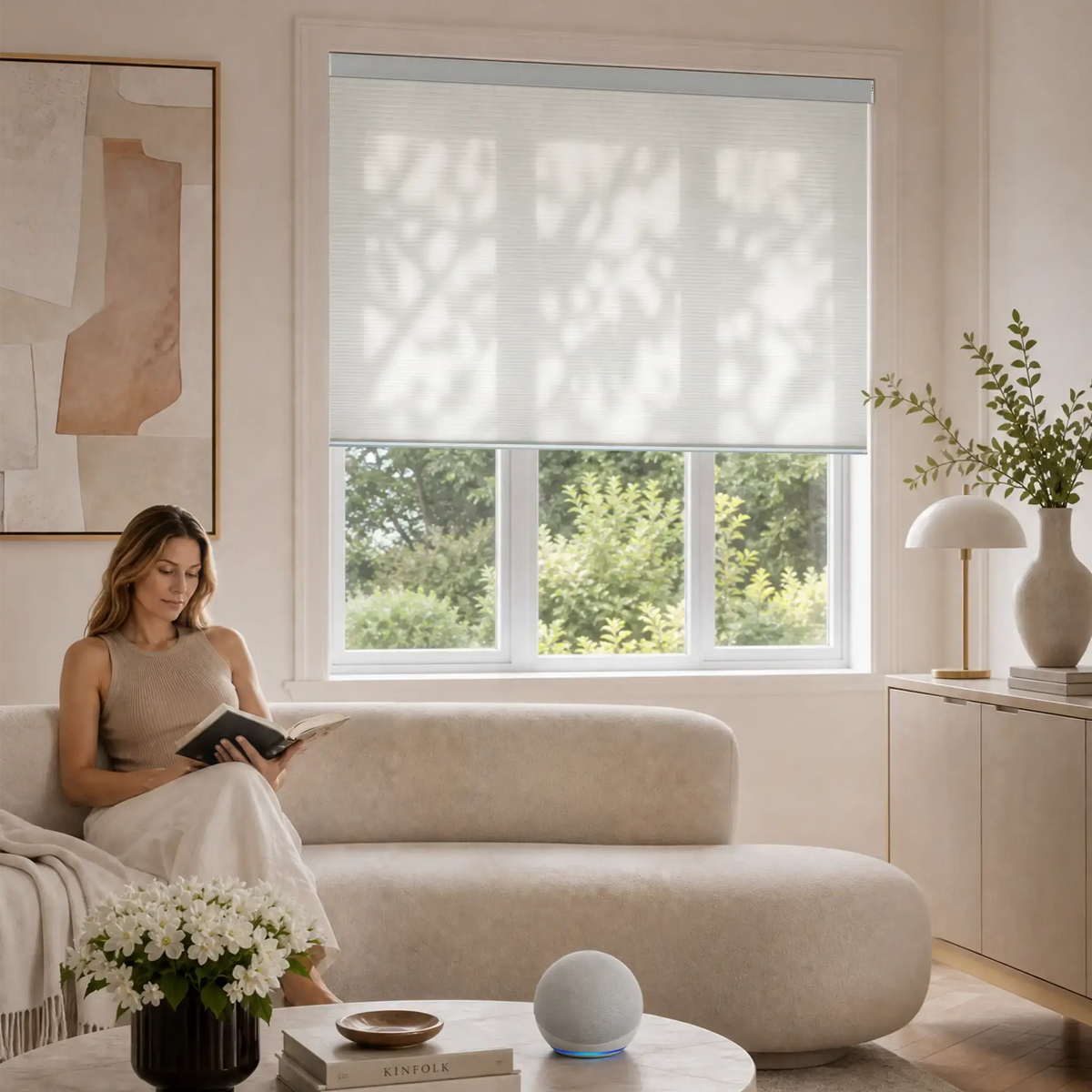 A woman sits on a beige sofa in a bright, modern living room, reading a book near a large window with amotorized cellular shades partially drawn. The room features neutral beige walls, a white sideboard with a lamp and vase, and a round coffee table with a small speaker and books.