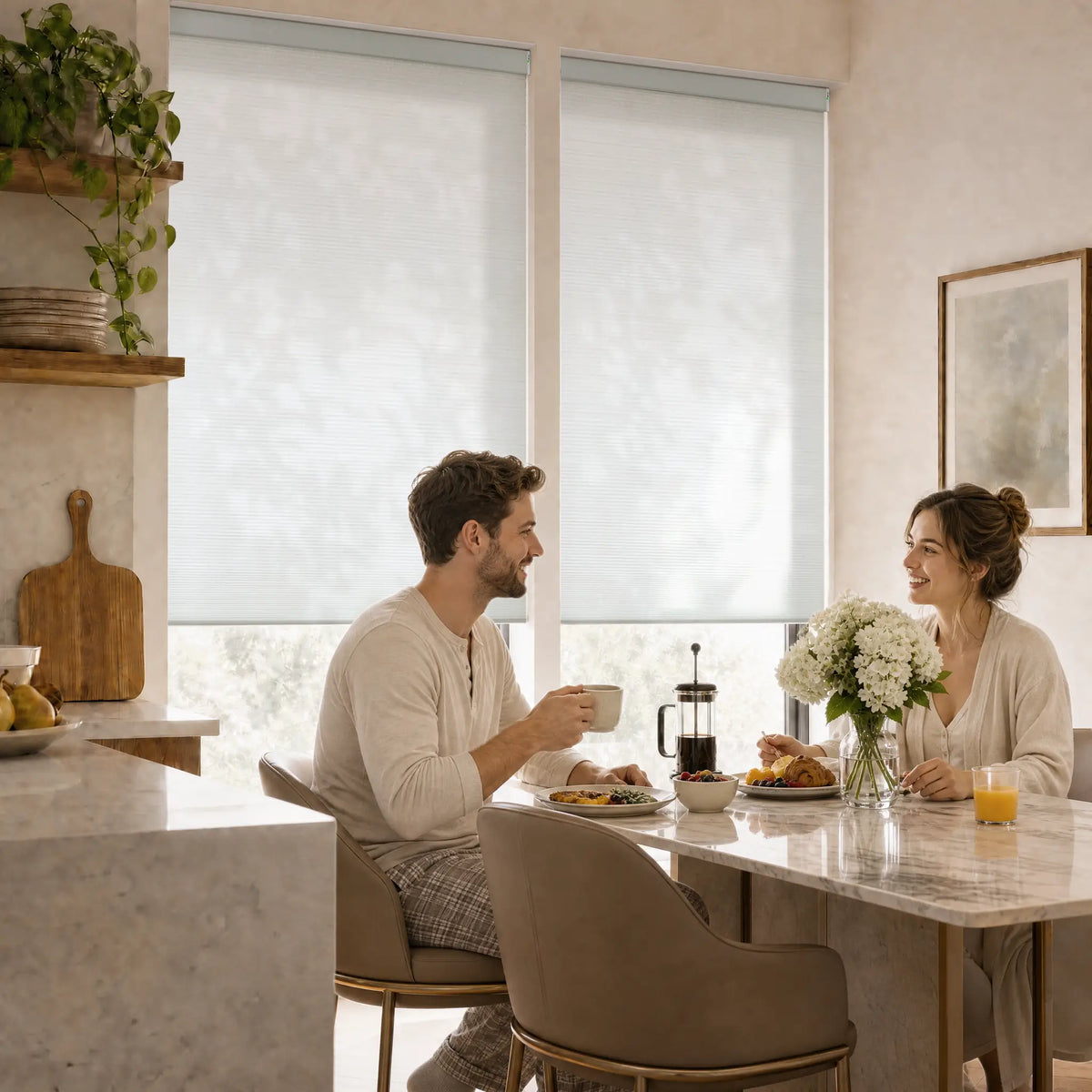 A man and a woman sit at a marble dining table in a bright, modern kitchen, sharing a light breakfast. The room features two large motorized cellular shades that diffuse natural light, creating a warm and inviting atmosphere.