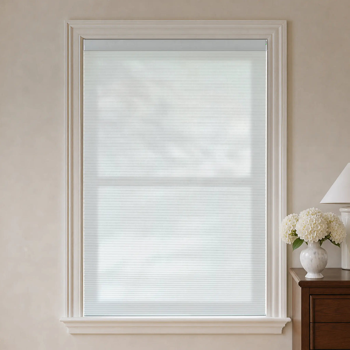 A white motorized cellular shade installed in a tall white-trimmed window, shown in a styled room setting with a wooden side table, lamp, and white flowers on the right.