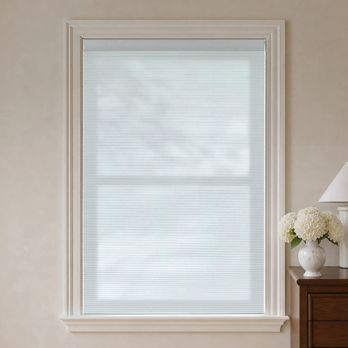 A white light filtering cellular window shade installed in a tall white-framed window, shown in a softly lit room with beige walls, a wooden side table, and a white vase of flowers.