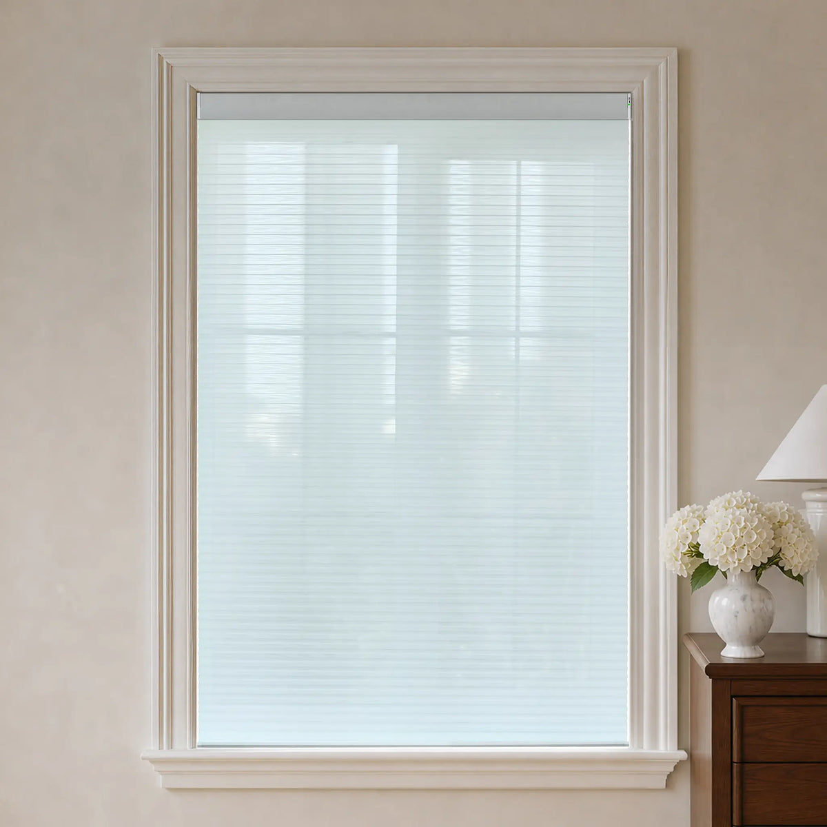 White light filtering cellular window shade installed in a white window frame, shown in a styled room setting with a wooden side table and a vase of white flowers on the right.